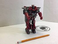 Red and gray transformer robot figure standing upright on a white surface next to a pencil for size reference