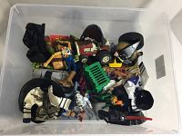 Top-down photo of a plastic bin containing a mixed assortment of toys including action figures, a toy vehicle, and accessories.