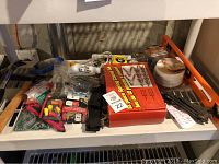 Photo shows a shelf with assorted hardware tools including a red box of a 12-piece spring clamp set, tape measures, a bright orange level, a hammer, and several pliers and hand tools.