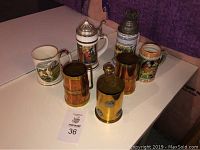 Six items on a table showing three German beer steins and three brass canisters, some with decorative lids and artwork.