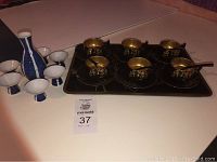 Full view showing blue and white ceramic sake set with carafe and four sake cups, alongside black tray holding six black and gold tea cups with saucers and spoons.