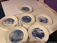 Set of 6 vintage Wedgwood winter plates laid out on a white surface showing blue and white transfer printed winter scenes with skiers and snowy landscapes.