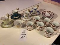 Full set of demitasse cups and saucers arranged in rows on table, showing multiple designs and presence of gold accented handles and floral patterns.
