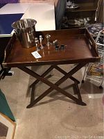 Wooden tray holding metal ice bucket and salt and pepper shakers on folding legs in basement setting