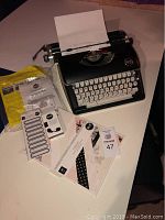 Black We R Typecast typewriter with sheet of white paper loaded, scrapbooking accessories laid out next to it including label stickers, templates, and instruction manuals.
