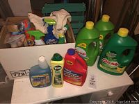 Photo of three large Cascade dishwashing liquid bottles and several other cleaning products packed on a table, including Woolite, Vim, carpet cleaner, and other spray bottles and cloths in a box.