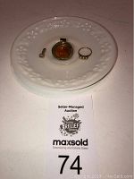 Three jewelry pieces displayed on white milk glass saucer along with auction lot number card.