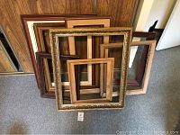 Photo showing multiple empty wooden picture frames stacked against wood-paneled wall on carpeted floor.