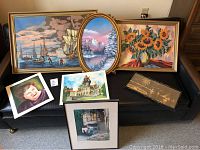 Eight various framed art pieces placed on a black bench, showing sailing ships, winter landscape, sunflowers, portrait and architectural prints.