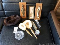 Various vintage items including mink hat, vanity brush and mirror set, glass bowl, carved figurines on wooden plaques, floral art, glass perfume bottle, decorative trinket box, and comb displayed on black cushioned surface.