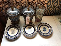 Photo showing two blue and white ceramic containers with lids on metal stands, six blue and white decorative plates, and one tall heavy glass vase with red glass bottom.