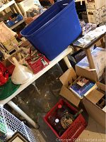 Wide view of wine making items including large blue plastic bin, carboys, corking machine, drying rack, boxes, and scattered tools.