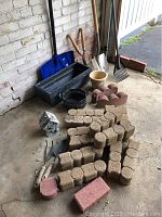 Assorted garden and landscape items including planter boxes, tools, and bricks arranged near a brick wall, showing tools like snow shovel and rake, plastics pots, and bricks in different shapes.