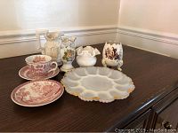 Wide shot of assorted vintage porcelain and milk glass items including teacups, jugs, vase, and scalloped plate on wooden surface.