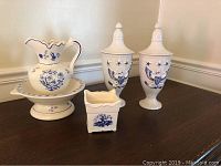Photo showing the two porcelain lidded urns, the pitcher, the wash basin, and small Delftware planter on a table with beige wall background.