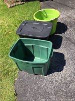 Three plastic containers outdoors on pavement and grass: dark green rectangular bin, black rectangular bin with lid, round green tub with rope handles.