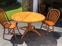 Photo of round pine drop-leaf table and two oak chairs placed outdoors with visible wood grain and knots on table surface.