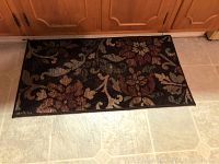 Closeup showing a 20" x 32" doormat with dark background and multicolored floral design underneath wooden cabinet.