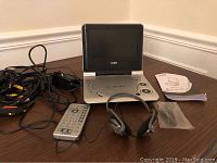 Full view of all items in the lot including the Coby portable DVD player open with screen, remote control, power cables, user manual, and a black headset sealed in plastic.