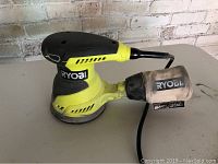 Ryobi orbital sander with dust catcher attached and cord visible on a table against a white brick wall.