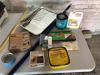 Lot of painting supplies laid on table with wood filler, primer container, paint tray, rollers, trim and peel-and-stick tile.