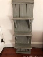 Front view of gray-green hanging wall shelf against wall showing three shelves and vertical slats, includes hanging hardware.