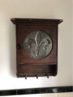 Front view of wooden hanging cabinet with opaque round glass door featuring fleur-de-lis design and three hooks at bottom.