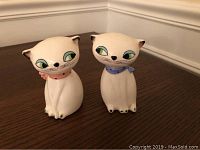 Front view of a pair of white cat-shaped salt and pepper shakers, one with a pink collar and one with a blue collar, ceramic with painted facial features and collars.