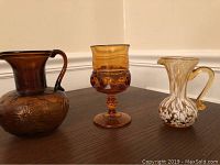 All three glassware pieces placed on a wooden surface showing relative size and color: amber thumbprint glass, amber depression glass pitcher, and spatter pitcher with white speckles.