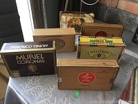 Six vintage cigar boxes displayed on a white surface with varying sizes and brands visible, including Muriel Coronas and House of Lords Queens