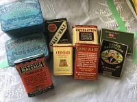 Nine assorted vintage tobacco and cigarette tins arranged on a white embroidered cloth, showing variety in brands and colors