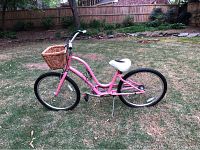 Side view of a pink cruiser bicycle by REI with a white seat and a brown woven front basket in a backyard.