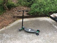 Black and green Fuzion scooter positioned on a concrete surface outdoors with greenery in background, showing general view and side profile.