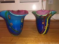 Front view showing both vintage blue glass art vessels with multicolored patterns and bright pink interior, placed on wooden floor with white background
