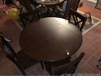 Photo showing round brown wooden table with visible marks, surrounded by four matching cross back chairs in a room with tiled flooring.