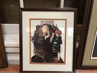 Framed Norman Rockwell print showing a doctor using a stethoscope on a doll held by a child, photographed indoors against a neutral background.