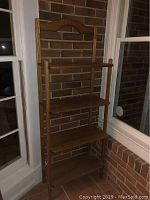 Front view of a tall brown wooden shelf with four shelves against brick wall between front window and door.