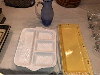 Photo showing white ceramic divided dish with textured grapevine pattern, yellow ceramic serving platter with floral/fruit design, and blue hand blown glass pitcher.