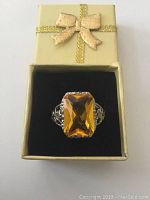 Silver tone ring with large amber colored rectangular gemstone in cream-colored box with gold glitter bow on top, shown from front.