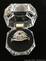 Close-up photo of one silver tone ring featuring a detailed gem-studded design inside a clear plastic box with black cushion