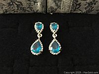 Close-up of silver tone drop earrings with blue teardrop gemstones and clear stone halos.