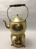 Front view of hammered brass tea kettle on warmer stand with wooden handle and curved spout.