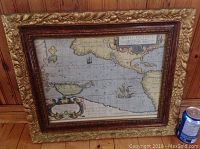 Full view of framed vintage map with ornate gold-colored gesso frame on wooden panel background with soda can for scale.