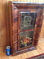 Front view of the large antique wood frame clock showing visible brass gears behind glass and floral panel below.