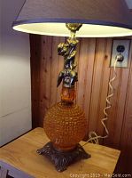 Full view of the lamp showing the amber hobnail glass body, brass cherub figure, metal base, fabric lampshade, and power cord
