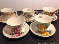 Five sets of vintage floral teacups and saucers arranged on black surface, showing varied floral designs and color schemes.
