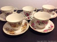 Photo showing five teacups and saucers with distinct floral designs and gold rims, arranged in two rows on a table.