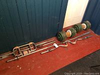 Photo showing two dumbbells with multiple green weight plates and various weight lifting bars placed on a reddish floor against a blue wall.