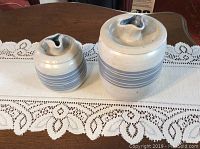 Front view of two matching pottery canisters on lace table runner, showing shape, lid design, and blue banding.