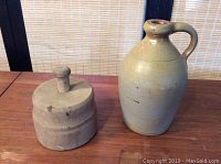 Photo showing small beige stoneware crock jug and wooden butter print stamp side by side on wooden table with bamboo backdrop.